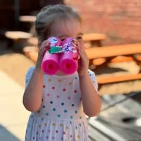 Child in a polka-dot dress looking through pink toy binoculars outdoors