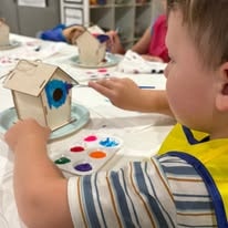 Young boy painting a wooden birdhouse at a craft table with a paint palette