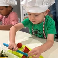 Child in chef hat and green shirt cutting tomatoes and vegetables on a cutting board