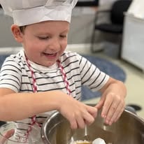 Smiling boy in chef hat and striped shirt stirring a large mixing bowl