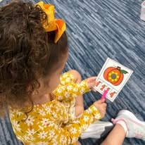 Child in yellow floral outfit playing with a pumpkin-themed counting card on a blue carpet