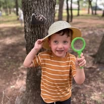Smiling boy in a sun hat holding a green magnifying glass by a tree