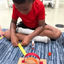 Child in a red shirt working on a paper craft with glue on a classroom carpet