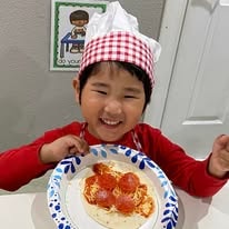 Smiling child in a chef hat holding a plate with a mini pizza