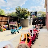 Outdoor play yard with ride-on toy cars, a playhouse, basketball hoop, and fences