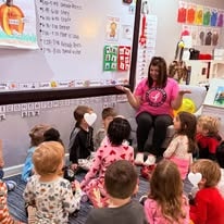 Teacher reading aloud to children seated on a classroom carpet in front of a smartboard