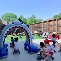 Children playing outside at a backyard party with a large inflatable shark-arch bounce house