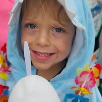 Child in a blue shark costume with flower leis smiling with a snow cone