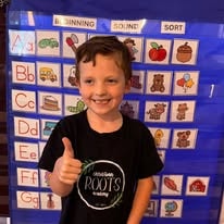 Smiling boy in Deep Roots t-shirt giving a thumbs-up in front of an alphabet phonics chart